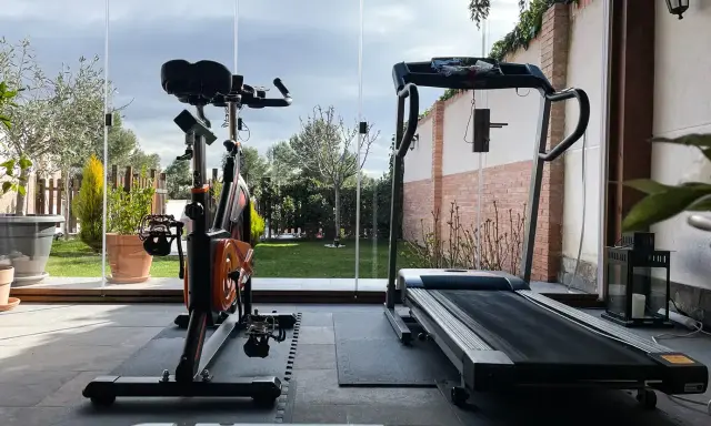 Un gimnasio en terraza con bicicleta estática y cinta de correr, con vistas a un jardín verde y un cielo nublado.