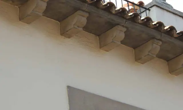 Detalle de qué es una cornisa, con ménsulas de piedra decorativas bajo tejas curvas y una barandilla metálica.