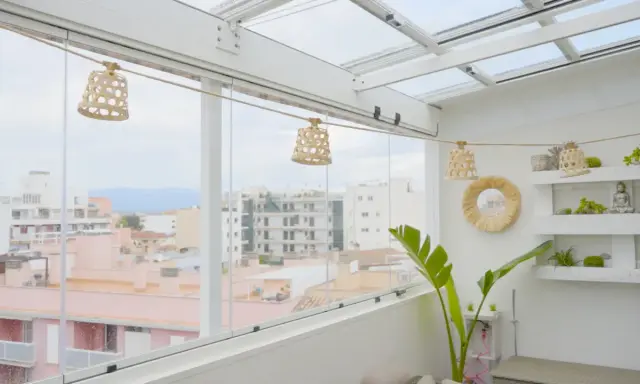 Un cerramiento ático con vistas urbanas, decorado con lámparas de mimbre y plantas.