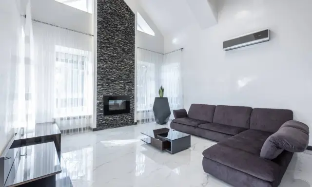 Un moderno salón con un sofá gris oscuro, chimenea de piedra y suelo de mármol blanco.
