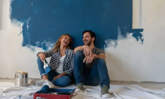 Pareja riendo sentada en el suelo frente a una pared recién pintada de azul. ¡Qué divertido es pintar una habitación!
