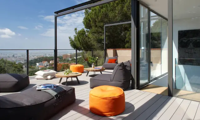 Cómodo balcón terraza con sillones, pufs naranjas y mesas bajas, con vistas a la ciudad.