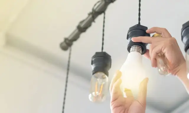Manos cambian una bombilla LED por una incandescente en una lámpara colgante, actualizando la luz del hogar.
