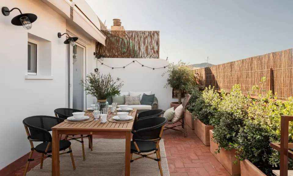 Acogedora terraza de 25 metros cuadrados con mesa de madera, sillas negras, sofá, plantas y luces decorativas.