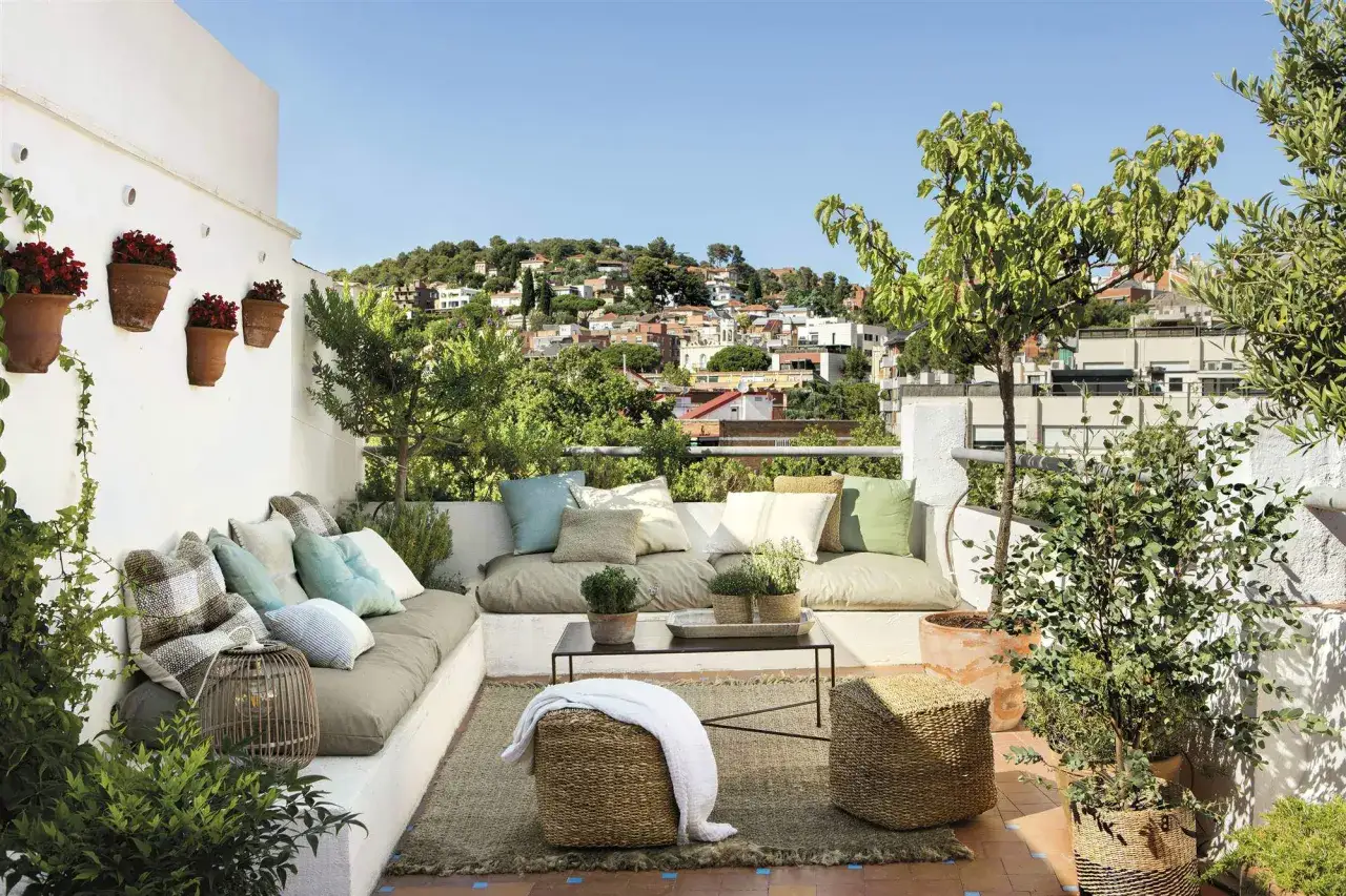 Acogedora zona chill out exterior con sof&aacute;s, cojines, alfombra y plantas. Un oasis de paz con vistas a la ciudad.