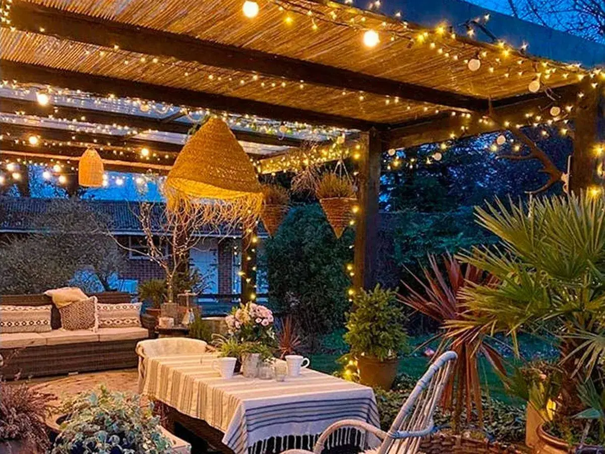 Un porche acogedor con luces cálidas, ideal para saber como iluminar un jardin. Muebles de exterior, plantas y una mesa puesta crean un ambiente mágico.