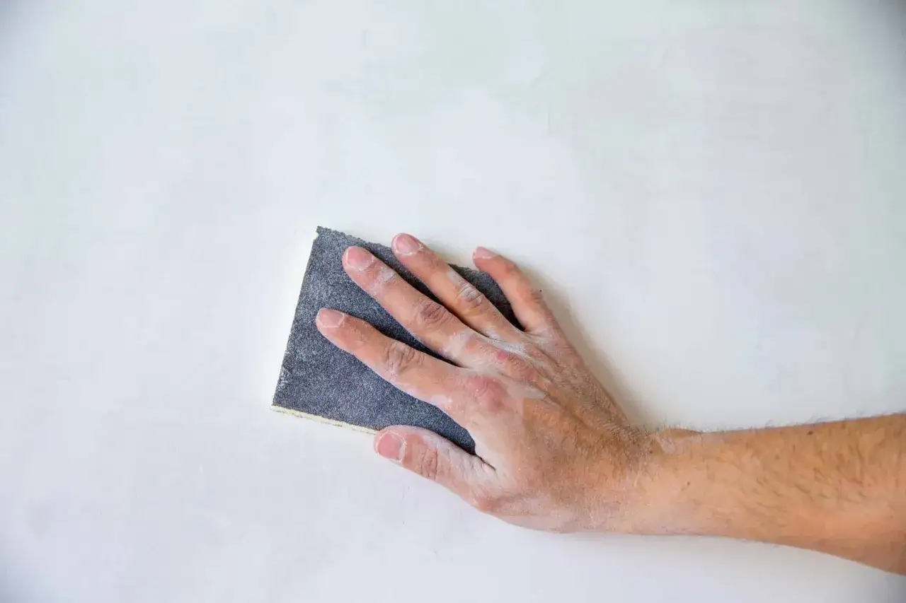 Mano lijando una pared blanca, paso esencial para saber como pintar una pared ya pintada de otro color y lograr un acabado perfecto.