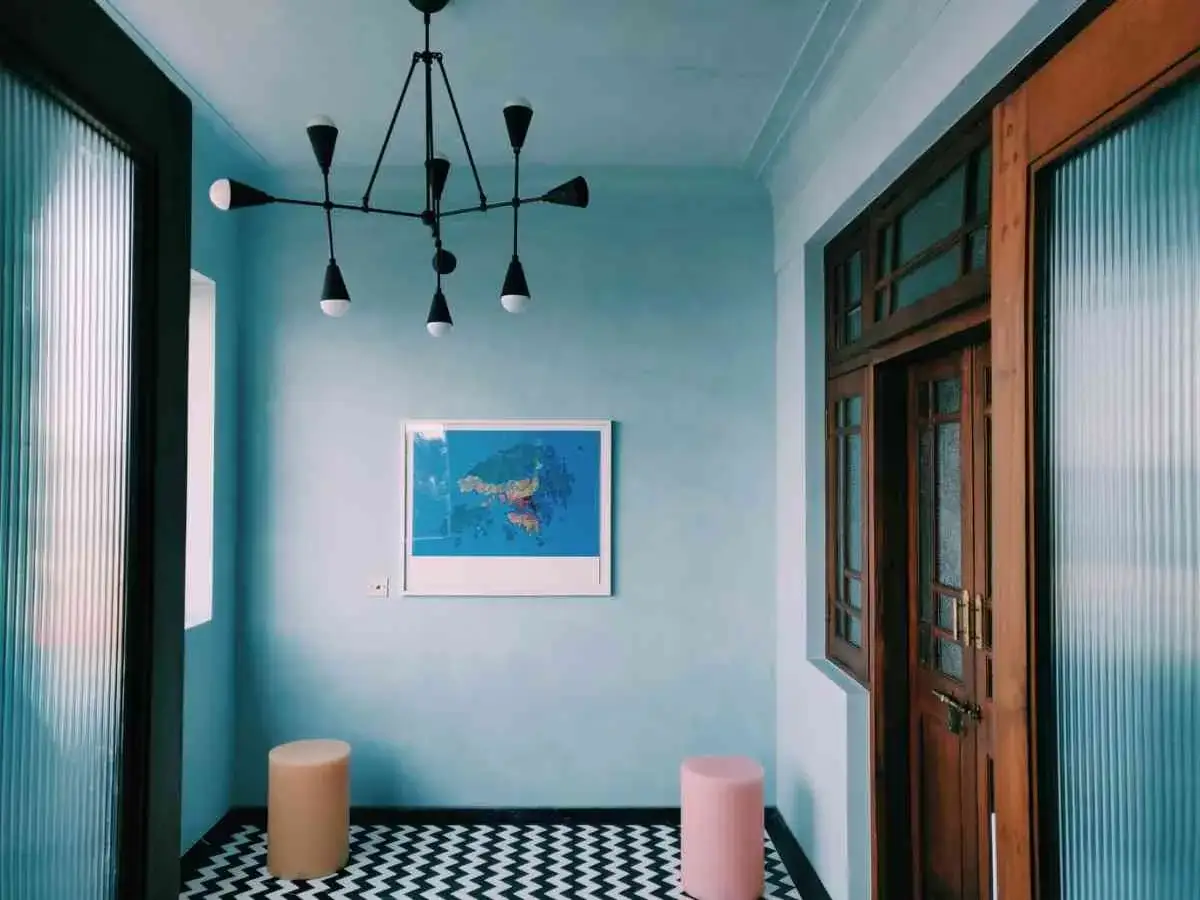 Un pasillo moderno con paredes y techos pintados de azul claro. El suelo tiene un patrón de zigzag blanco y negro. Hay un mapa colgado en la pared y dos taburetes redondos.