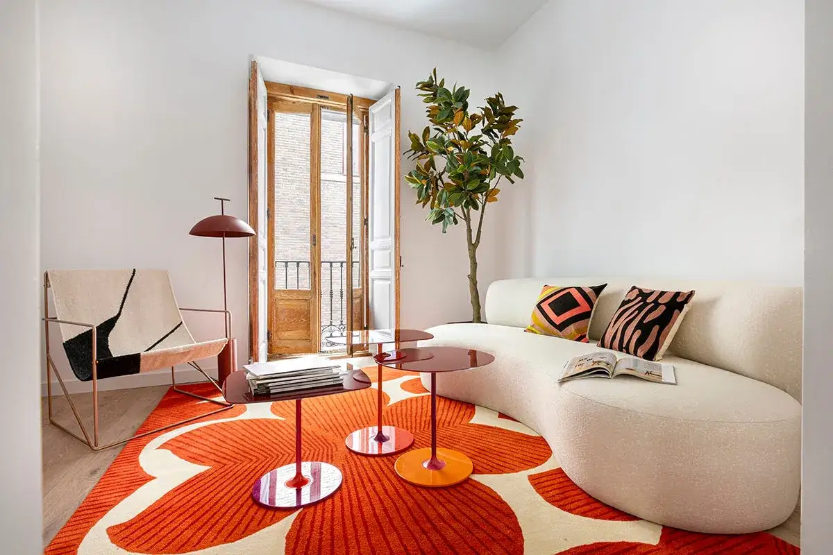 Sofá curvo blanco, alfombra naranja con flores, sillón moderno y mesa auxiliar, combinando diversos estilos de diseño.