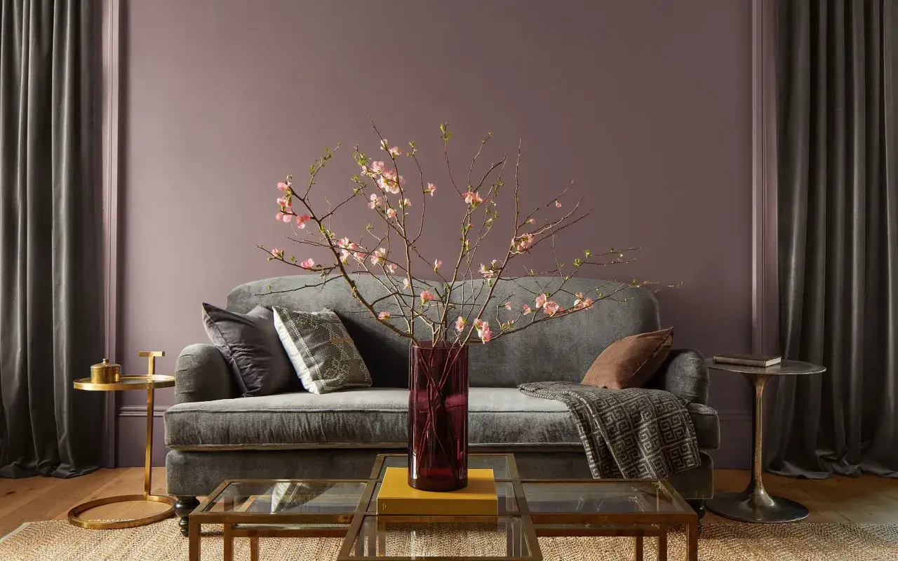 Sofá gris con cojines y manta, jarrón con flores rosadas, mesas doradas y de cristal. Un ambiente de estilo romántico diseño de interiores.