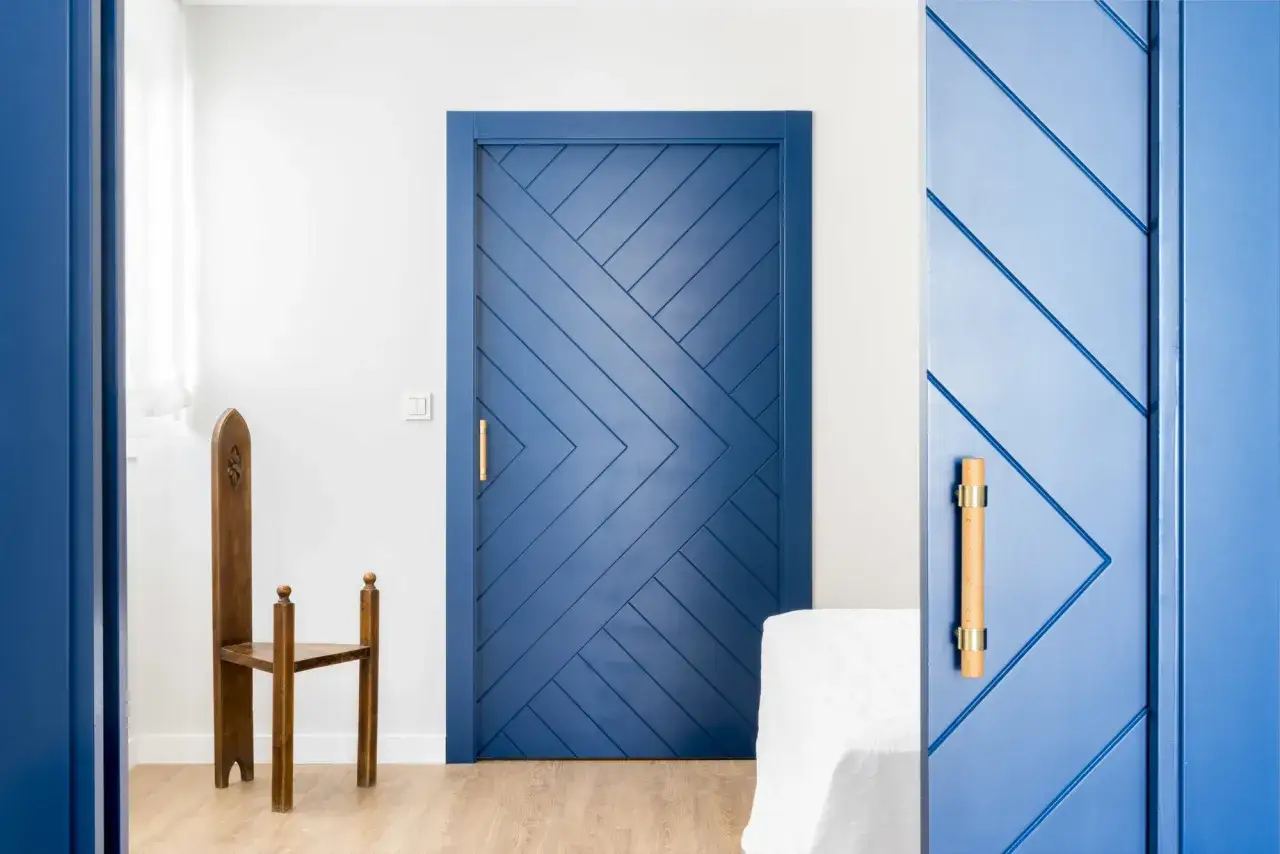 Puertas de dos colores, una azul con diseño de espiga y otra azul marino. Un sillón de madera antiguo y una mesa cubierta con tela blanca.