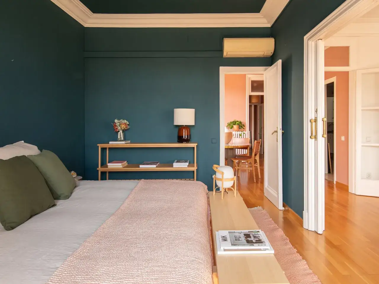Habitación con paredes azul oscuro y techos pintados originales con molduras blancas. Cama con mantas y cojines verdes. A través de puertas abiertas, se vislumbra un comedor con paredes melocotón.