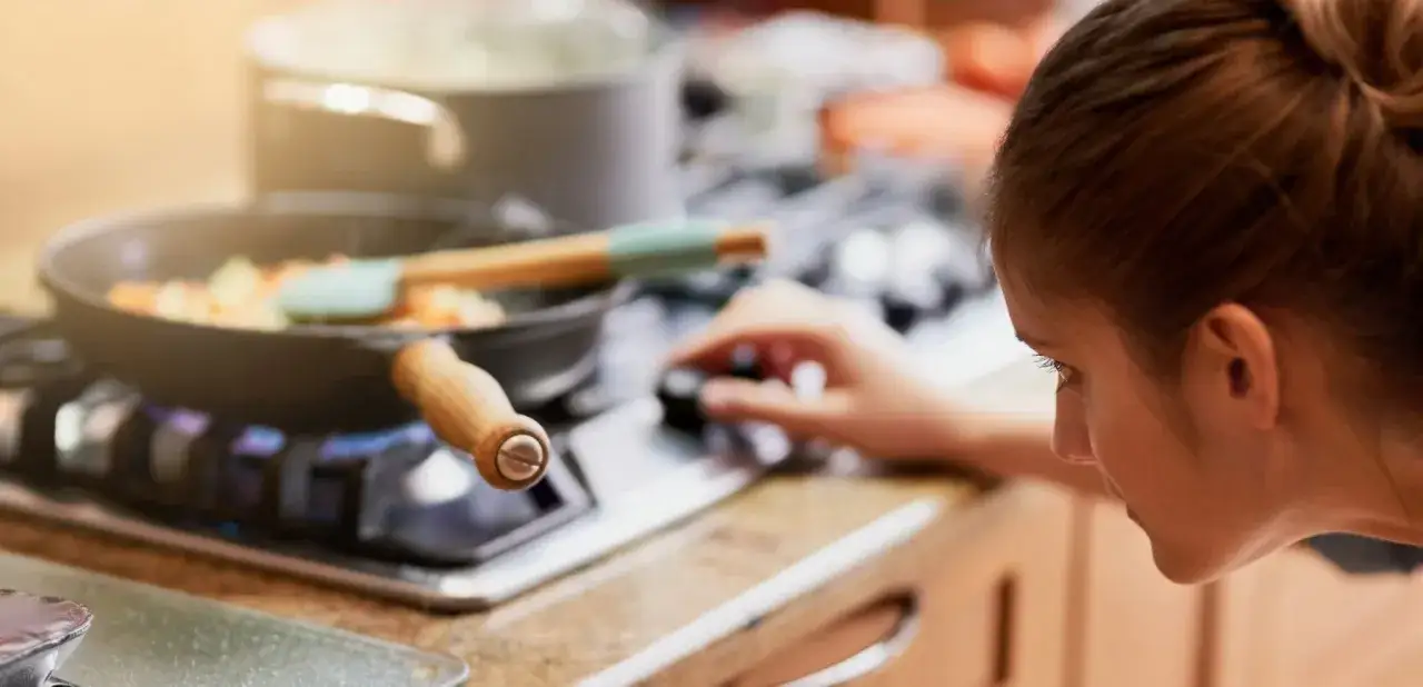 Mujer ajustando la perilla de la estufa de gas, con una sartén humeante encima. Cocinando con butano o gas natural.