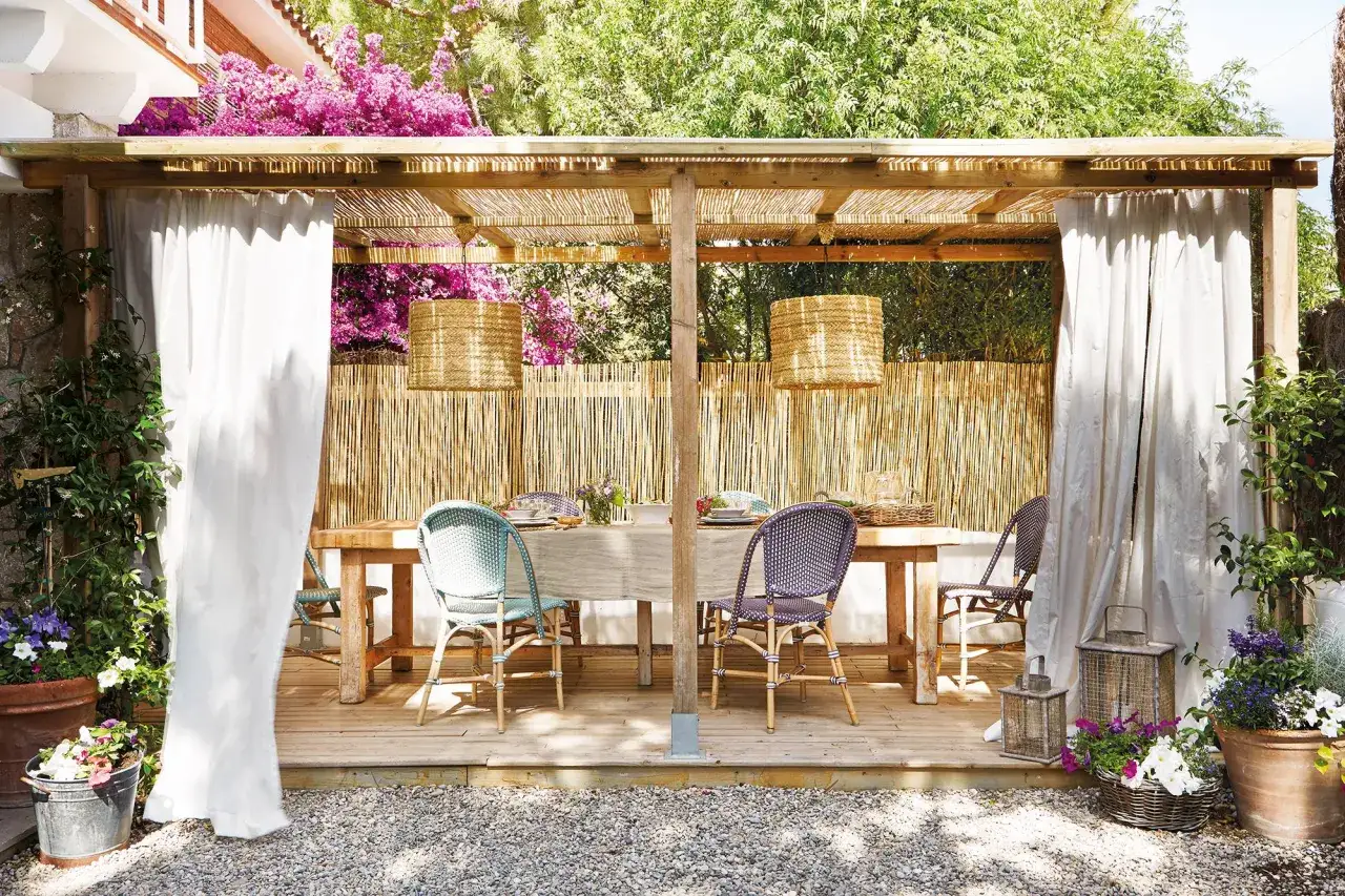 P&eacute;rgola de madera con enredaderas para pergolas, cortinas blancas y mesa puesta para comer al aire libre.