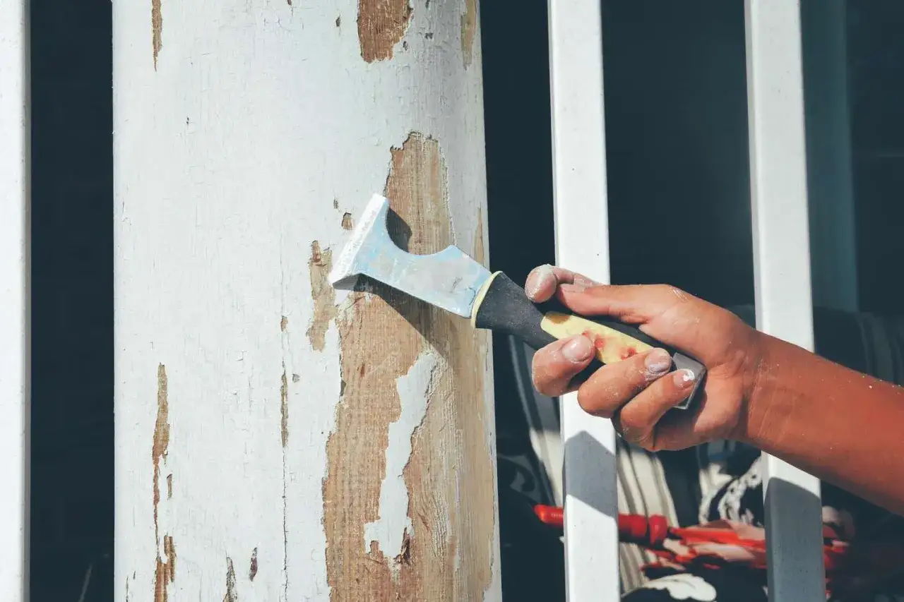 Mano raspando pintura descascarada de una columna de madera rugosa. Proceso de cómo quitar pintura de una pared rugosa.