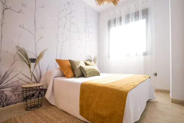Habitación con papel tapiz botánico y cama con cojines naranjas y verdes. Una manta mostaza ilumina el espacio, ideal para quienes buscan colores para paredes con gotele.