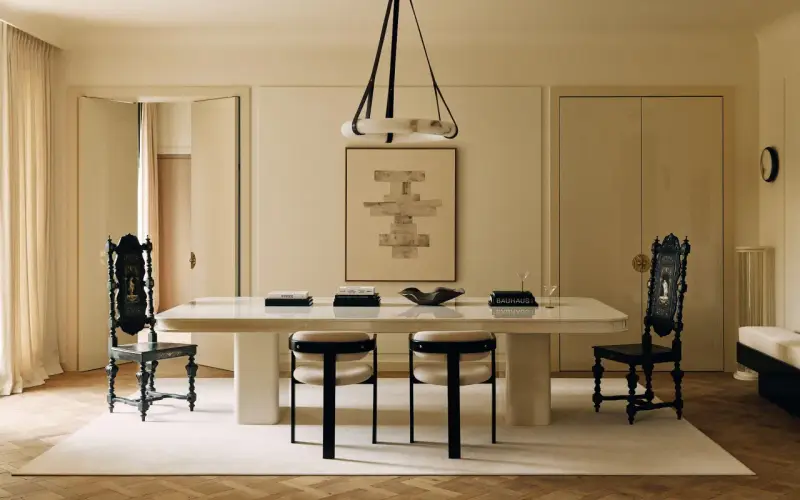 Comedor elegante con mesa blanca, sillas negras y una paleta de colores crema. Arte abstracto y cortinas suaves complementan el espacio.