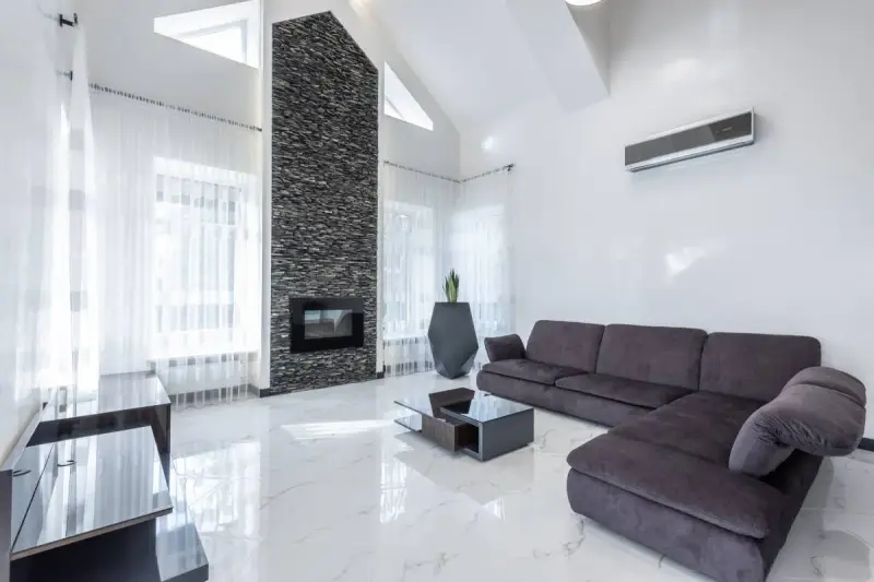 Un moderno salón con un sofá gris oscuro, chimenea de piedra y suelo de mármol blanco.